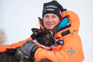 Appetitt ambassadør og hundekjører Marit Beate Kasin holder sin sledehund tett inntil seg, hun er ikledd tykk oransje vinterjakke.