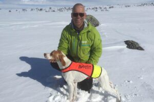 Appetitt ambassadør Rune Hoholm med grønn Appetitt-jakke smiler til kameraet, sittende i snøen sammen med sin fuglehund iført vest fra Appetitt.
